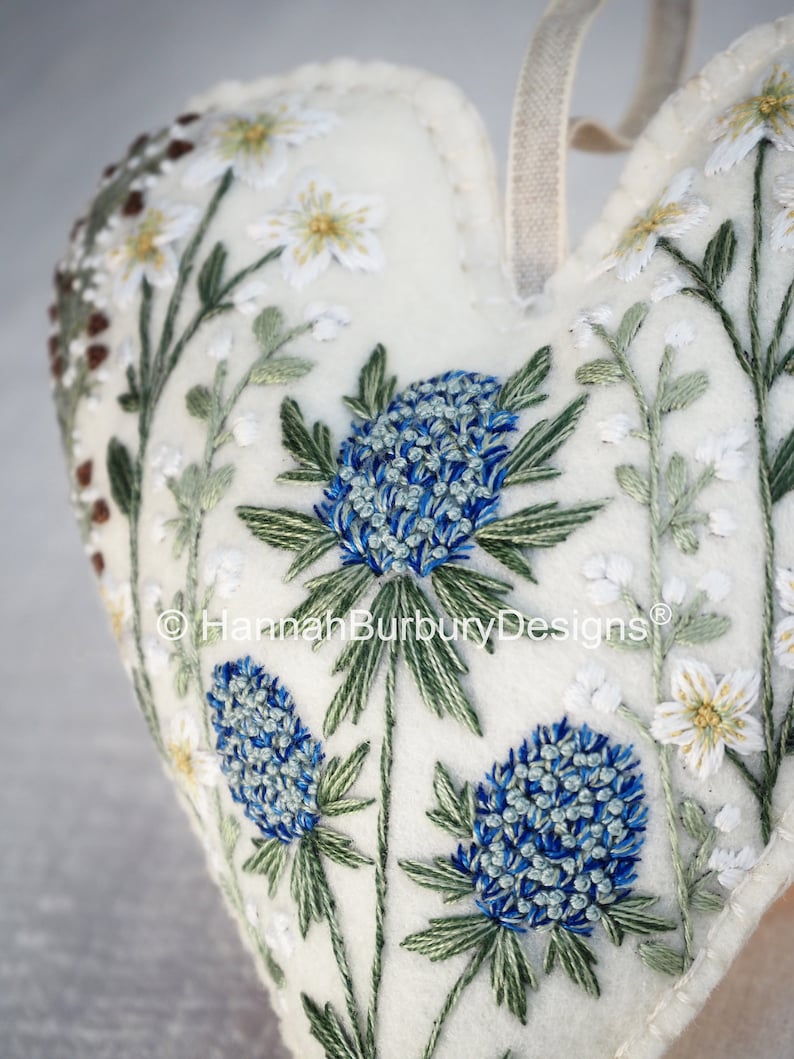 May include: White felt heart-shaped ornament with blue and green embroidered flowers. The ornament has a white ribbon loop for hanging. The text "Hannah Burbury Designs" is embroidered on the ornament.