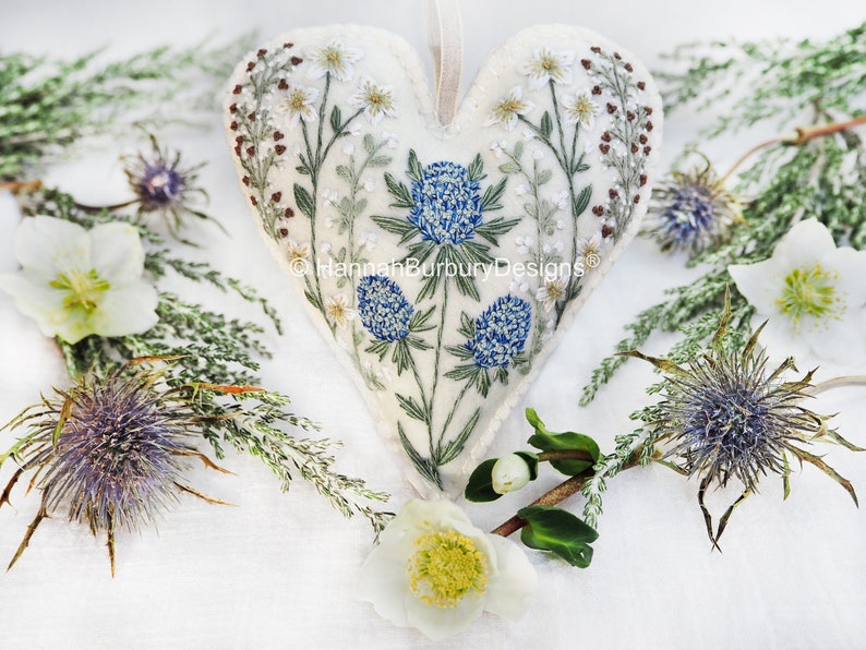May include: A white felt heart-shaped ornament with blue and white embroidered flowers. The ornament is surrounded by white flowers and green foliage.