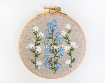 May include: Embroidered hoop art featuring blue forget-me-nots and white daisies on a beige linen background. The hoop is made of wood and has a brass screw closure. The text "Hannah Burbury Designs" is embroidered in the lower centre of the hoop.