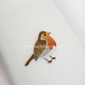 May include: A white, folded fabric with an embroidered robin. The bird features brown, orange, and white thread detailing. The robin is perched on the fabric, with its legs and beak visible. The text "HannahBurburyDesigns" is in the upper left corner.