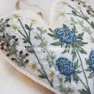 May include: A white felt heart-shaped ornament with embroidered blue and green flowers and white flowers with yellow centres. The ornament has a loop for hanging and the text "HannahBurburyDesigns&reg;" is embroidered on the front.
