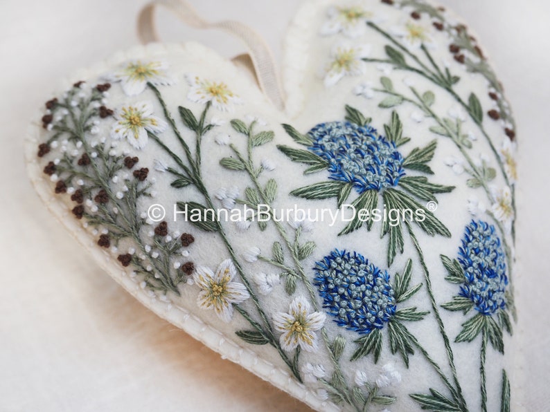 May include: A white felt heart-shaped ornament with embroidered blue and green flowers and white flowers with yellow centres. The ornament has a loop for hanging and the text "HannahBurburyDesigns&reg;" is embroidered on the front.