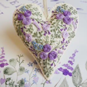 May include: A heart-shaped ornament made of off-white felt featuring intricate hand embroidery of purple roses, small blue flowers, and green foliage. A light-colored ribbon loop is attached to the top. The text HannahBurburyDesigns is visible.
