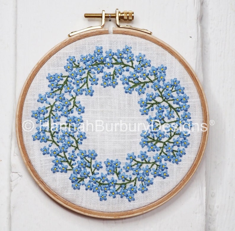 May include: A blue and green embroidered wreath of forget-me-not flowers in a wooden embroidery hoop. The hoop is on a white background.