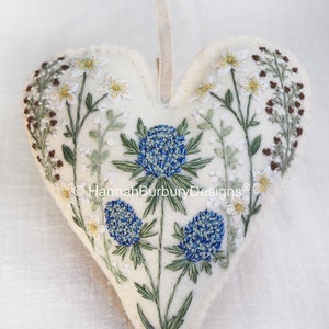 May include: A white felt heart-shaped ornament with blue and green embroidered flowers. The ornament has a loop for hanging and the text "&copy; Hannah Burbury Designs&reg;" is embroidered on the bottom.