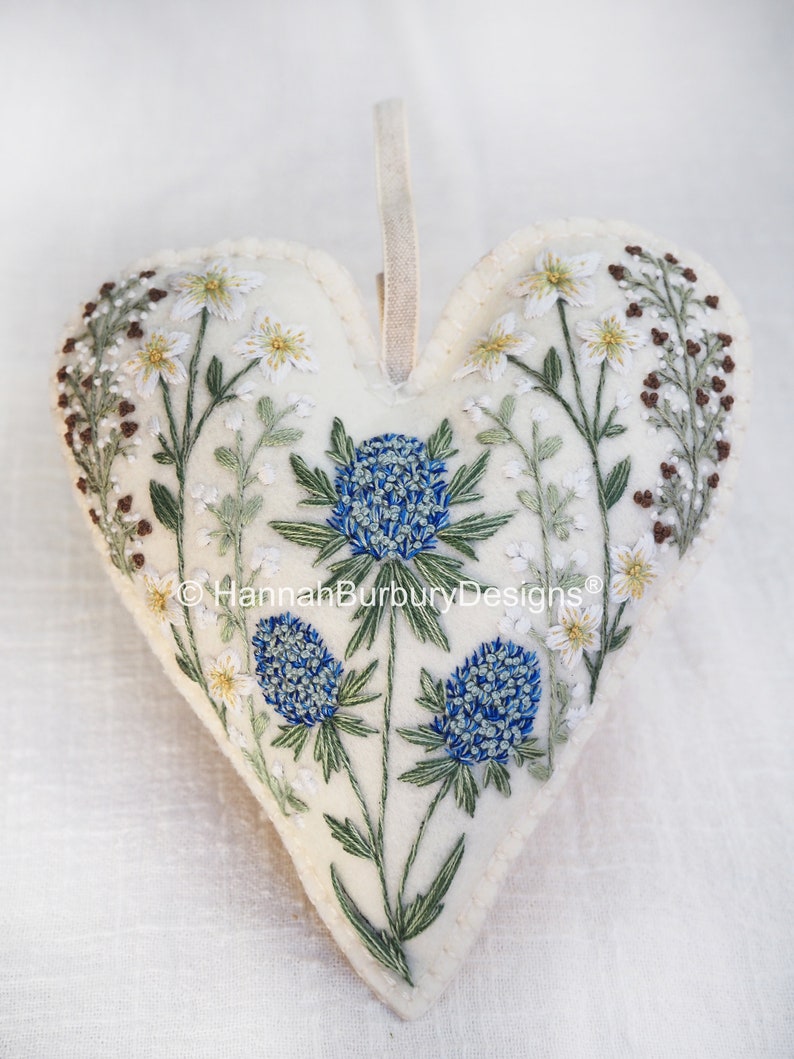 May include: A white felt heart-shaped ornament with blue and green embroidered flowers. The ornament has a loop for hanging and the text "&copy; Hannah Burbury Designs&reg;" is embroidered on the bottom.