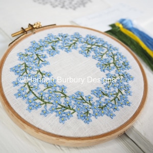 May include: A close-up of a completed embroidery hoop with a wreath of forget-me-not flowers. The hoop is made of wood and has a white linen fabric stretched over it. The flowers are embroidered in shades of blue and green.