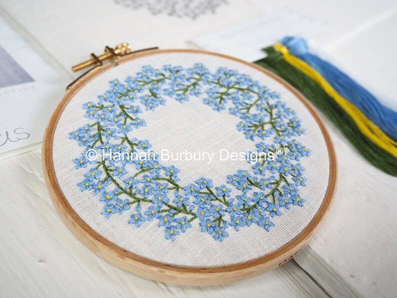 May include: A close-up of a completed embroidery hoop with a wreath of forget-me-not flowers. The hoop is made of wood and has a white linen fabric stretched over it. The flowers are embroidered in shades of blue and green.
