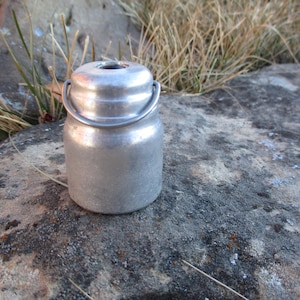 May include: A small, vintage, silver-colored metal container with a lid and a handle. The container is cylindrical with a rounded top and bottom. The background is a natural setting with rocks and dry grass.