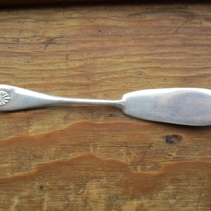 May include: A silver butter knife with a decorative floral design on the handle. The blade is wide and rounded. The knife is set against a wooden background.