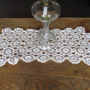 May include: White crocheted doily with a delicate pattern, featuring a clear glass oil lamp with a white wick in the center.