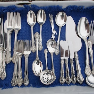 May include: An assortment of antique silver-plated flatware, including knives, forks, spoons, and serving utensils, arranged on a blue cloth inside a white tray. The silverware features ornate, detailed handle designs.