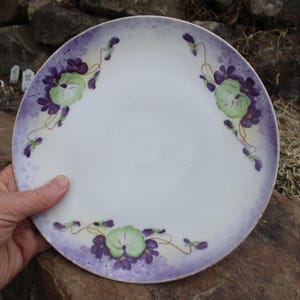 May include: A white porcelain plate with a purple and green floral design. The plate features a delicate pattern of pansies with purple petals and green leaves.