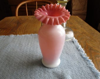 May include: A white and pink glass vase with a ruffled pink top. The vase is sitting on a blue and white textured surface.