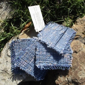 May include: A set of square, woven fabric coasters in shades of blue and white with frayed edges. A white rectangular tag is attached to the coasters, featuring the printed text "Mushroom Mug Rugs" along with additional descriptive information.