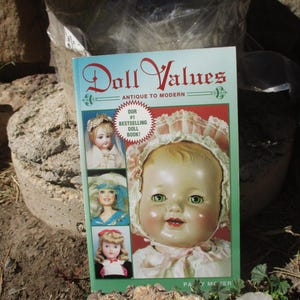 May include: A book titled "Doll Values" with the subtitle "Antique to Modern" is displayed. The cover features a large doll with rosy cheeks and green eyes, along with three smaller doll images. The book is labeled "Our #1 Bestselling Doll Book!"