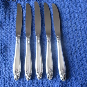 May include: Five silver knives with ornate floral detailing on the handles are arranged on a blue textured cloth. The knives have a classic design, with a shiny, reflective surface.