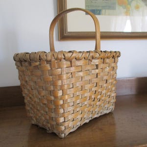 May include: A woven basket with a natural wood finish and a single handle. The basket is made of a light brown material and has a rectangular shape.