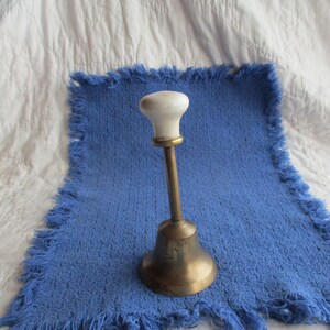 May include: A brass bell with a white ceramic knob on top. The bell is sitting on a blue fringed surface.