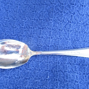 May include: A silver sugar spoon with an ornate design on the handle. The spoon is on a blue fabric background.