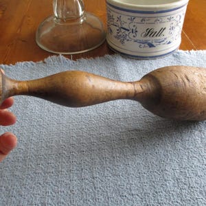 May include: A vintage wooden muddler with a bulbous head and a slender handle. The muddler is a warm brown color and appears to be well-worn. It is resting on a blue cloth, with a salt cellar and glass object in the background.