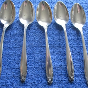May include: Five silver spoons with elongated bowls and detailed floral designs on the handles. The spoons are arranged on a blue textured surface, reflecting light. The spoons are likely for serving or dining.