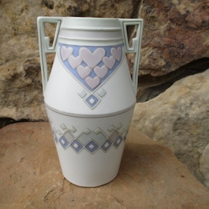 May include: A white ceramic vase with two handles and a geometric design featuring pink and blue hearts. The vase has a white background with a blue and gray geometric pattern.