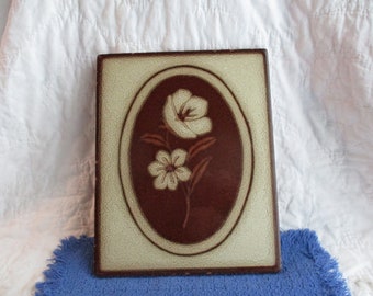 May include: A ceramic tile with a brown oval frame and a white floral design. The flowers are stylized and have a simple, elegant look.