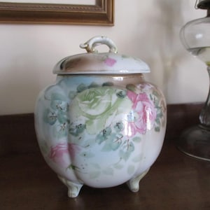 May include: A white porcelain cookie jar with a lid and three feet. The jar is decorated with hand-painted pink and green roses.