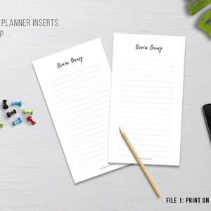 May include: Two white lined "Brain Dump" printable planner inserts with the text "Brain Dump" in a handwritten font. A pencil, colorful push pins, a smartphone, and a small potted plant are also visible.