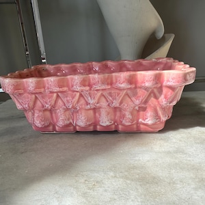May include: A rectangular, pink ceramic planter with a textured, geometric pattern. The planter has a scalloped rim and a glossy finish with white accents. The overall design is vintage and decorative, suitable for indoor or outdoor use.