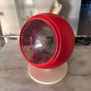 May include: A spherical red table lamp with a circular off-white base. The front features a clear, faceted glass lens covering an internal light bulb. A black power cord extends from the back of the red housing, which is mounted on the base.