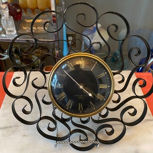 May include: A black metal clock stand with a gold clock face. The clock face has black Roman numerals and a black hand. The clock stand is shaped like a fan with intricate swirls.