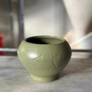 Puede incluir: Un pequeño jarrón redondo de cerámica verde salvia. El jarrón tiene una abertura ancha y un cuerpo redondeado con una base ligeramente acampanada. Dos diseños en forma de hoja están grabados en el lateral. El jarrón está sobre una superficie de mármol.
