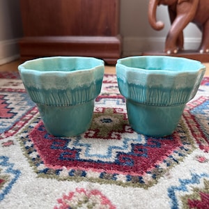 May include: Two matching ceramic planters with a scalloped rim and a glossy turquoise glaze. The upper section features vertical ribbed detailing, while the lower portion is smooth. Both vessels have a tapered shape and a consistent teal color.