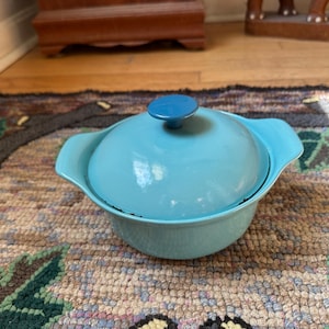 May include: A light blue, lidded casserole dish with a matching blue knob. The dish has two handles and sits on a patterned rug. The dish is likely made of ceramic or enamel and is suitable for cooking and serving.
