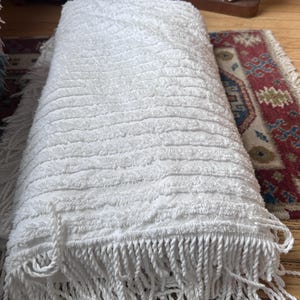 May include: A rectangular white textile throw blanket featuring a textured surface with horizontal raised stripes and long, twisted fringe detailing along the edges. The fabric has a soft, looped pile construction throughout the entire piece.