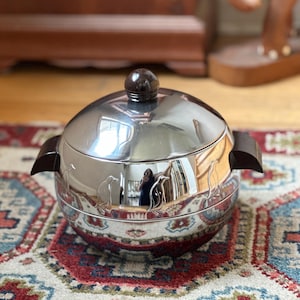 May include: A polished chrome ice bucket with a rounded lid and dark brown handles. The exterior features a series of embossed penguin silhouettes encircling the middle section. A small, spherical dark brown knob sits atop the lid.
