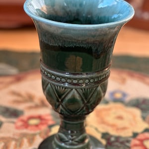 May include: A tall, green ceramic goblet with a flared rim and a decorative diamond pattern. The goblet has a pedestal base and a two-tone glaze, with a lighter blue-green color at the rim and a darker green on the body. The goblet is likely used for decorative purposes.