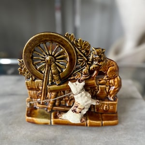 May include: A ceramic figurine depicting a spinning wheel scene with a dog and a cat. The figurine is primarily brown and tan, with intricate details. The dog is white with brown spots, and the cat is brown.