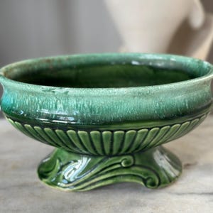May include: A green ceramic bowl with a pedestal base. The bowl has a wide, shallow design with a textured, glossy finish. The base features a fluted pattern and decorative scrollwork. The color is a vibrant, shaded green.