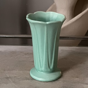 May include: A small, light green ceramic vase with a scalloped rim and vertical fluted detailing along the body. The vase features a flared opening that tapers down to a circular base, showcasing a matte finish and consistent color throughout.