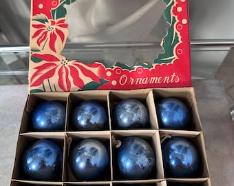 Pode incluir: Um conjunto de doze enfeites de Natal redondos de vidro azul-escuro com um acabamento desgastado. Os enfeites estão dispostos numa caixa de cartão com uma borda vermelha, ilustrações de poinsétias e o texto Christmas Tree Ornaments.