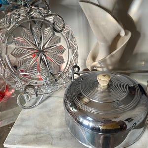 May include: A clear, cut-glass serving dish with a starburst design, displayed on a metal stand. Beside it is a chrome-plated, round serving dish with a cream-colored knob on the lid. The items are on a marble surface.