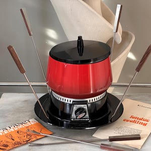 May include: A red and black electric fondue pot with a black lid, a temperature control dial labeled Cornwall, and four metal fondue forks with wooden handles. Includes two paper booklets titled Fun 'N Fancy Fondue Feasts and Fondue Cooking.