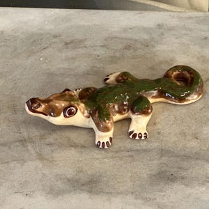 May include: A ceramic figurine of a crocodile with a glossy finish. The reptile is colored in shades of cream, brown, and dark green, featuring a long tail that curves to the side, four short legs with visible toes, and a detailed head.
