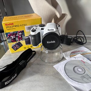 May include: A white Kodak PIXPRO AZ361 digital camera with a black lens cap, accompanied by its original yellow box, a black branded neck strap, a power adapter, and two software discs labeled with the camera model and user manual information.