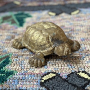 May include: A small, antique-style turtle figurine in a bronze finish. The turtle has a textured shell and detailed legs and head. The figurine is resting on a multi-colored, patterned rug.