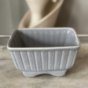 May include: A small, square, light blue ceramic dish with vertical ribbed detailing. The dish has a footed base and is likely vintage. The background is blurred, showing other ceramic pieces.