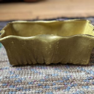 May include: A rectangular, gold-colored ceramic dish with a wavy rim. The dish has a slightly textured surface and is sitting on a patterned rug. The dish is empty and appears to be vintage.